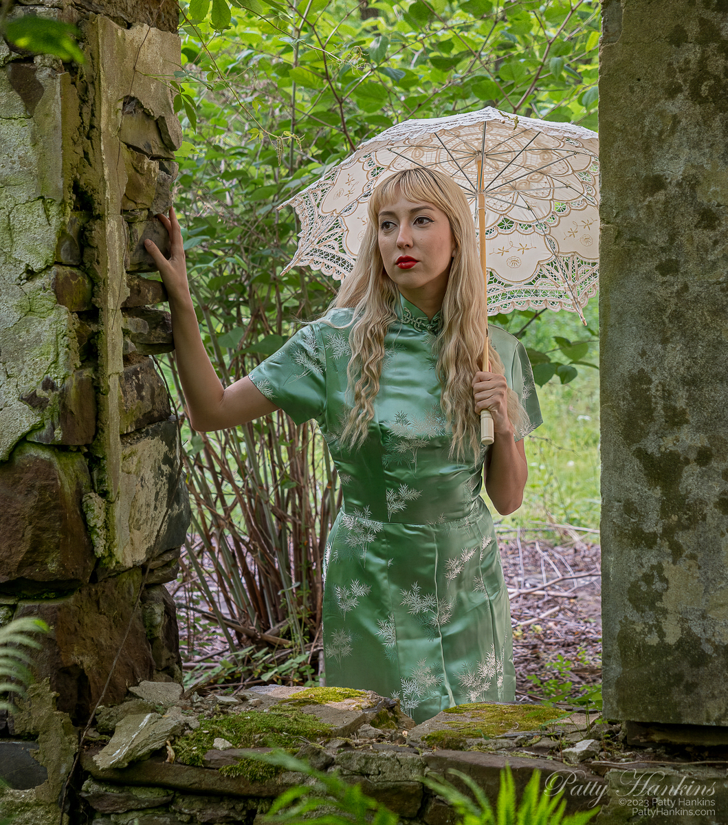 Lucy Magdlene, a white woman with long blond hair, wearing a 1960s era chinese inspired dress, holding an ivory parasol. She is standing in the ruins of a barn 