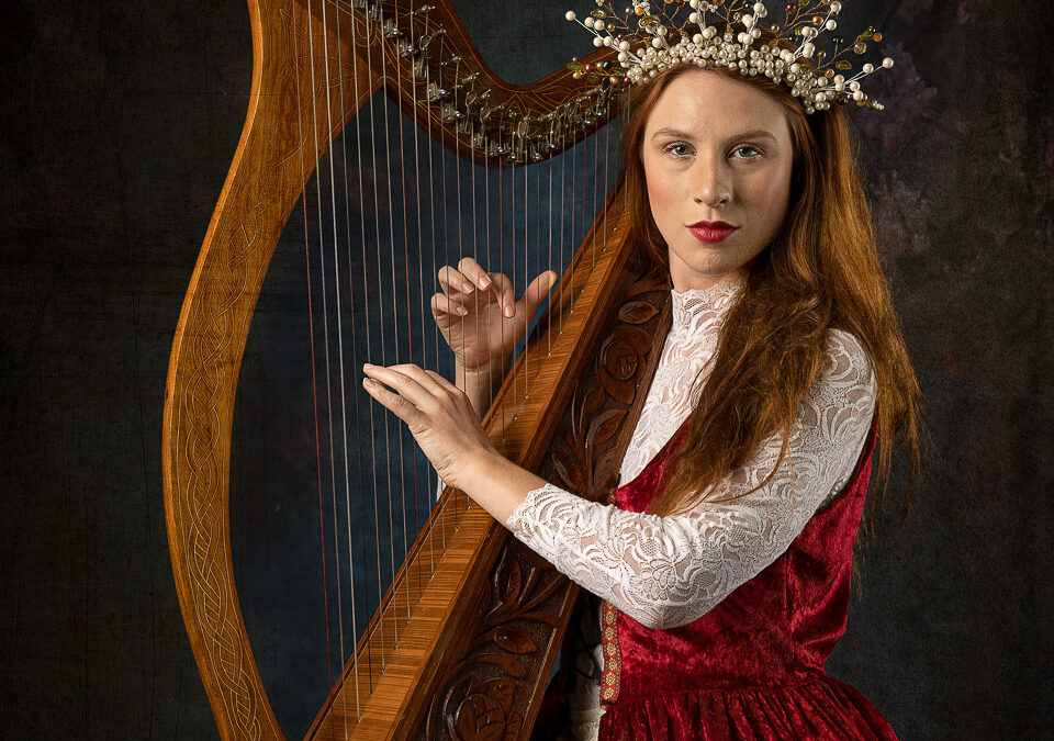 Astrid Kallsen wearing a renaissance inspired red over dress - over a white lace dress, wearing a pearl headpiece - playing her harp