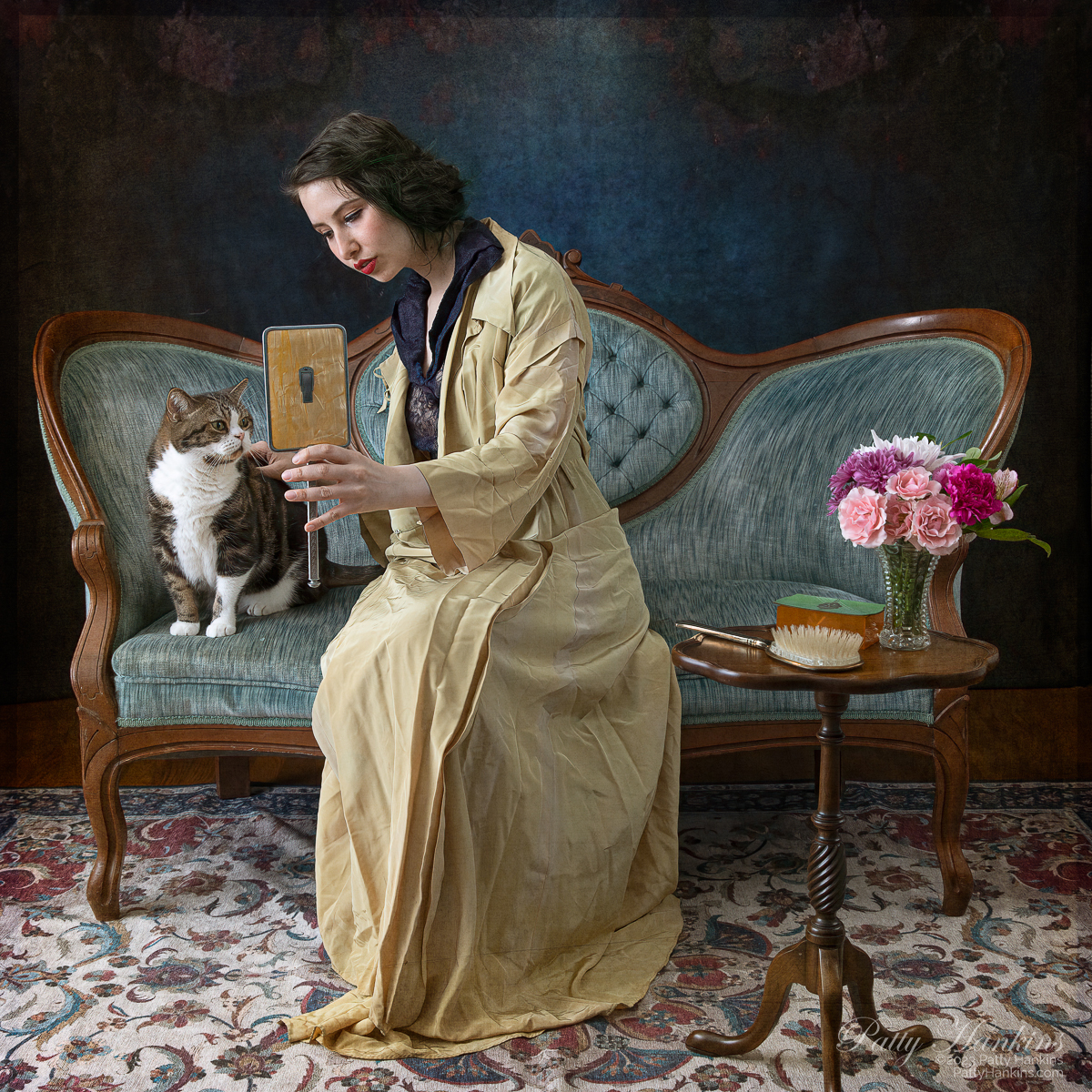 Woman sitting on a vintage sette holding a hand mirror so a brown and white tabby cat can look at himself in the mirror